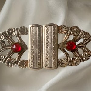 May include: An antique silver-toned clasp with ornate filigree detailing. The clasp features two rectangular center pieces and two red, faceted gemstones. The clasp is set against a white background.