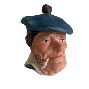 Puede incluir: Figura de cerámica de la cabeza de un hombre, con boina azul y un pequeño pompón marrón. La cara está pintada con tonos de piel realistas, un cuello blanco y un pequeño objeto en la boca.