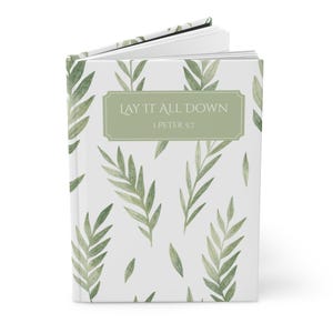 May include: A hardcover journal with a white background and a pattern of green leaves. The cover features the text "LAY IT ALL DOWN" and "1 PETER 5:7" in a green rectangular frame. The journal is open to reveal blank pages.