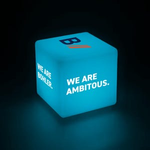 以下が含まれることがあります： 「WE ARE BOHLER.」と「WE ARE AMBITIOUS.」の文字が白で書かれた、光る水色の立方体。立方体の上面には、オレンジ色のストライプが入ったスタイリッシュな「B」ロゴがあります。暗い背景の中で立方体が照らされています。
