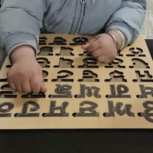 Peut inclure: Un plateau de puzzle carré en bois présentant une grille de découpes en forme de caractères de l'écriture devanagari. Le plateau est en bois clair et les pièces individuelles ont une finition sombre, texturée et à motifs dans les fentes.