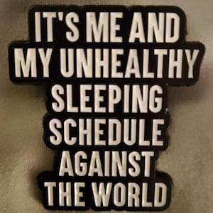 May include: A black rectangular enamel pin with raised white lettering that reads IT'S ME AND MY UNHEALTHY SLEEPING SCHEDULE AGAINST THE WORLD. The pin features a bold, block-style font and a smooth, matte finish with a distinct border around the text.