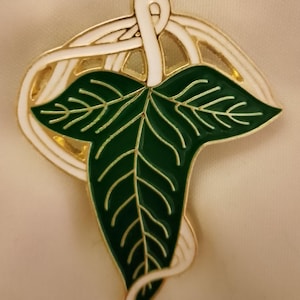May include: A gold-toned metal pin featuring a stylized green leaf with white and gold accents. The leaf has detailed veining and is set against a white and gold circular design. The pin is likely a decorative accessory.
