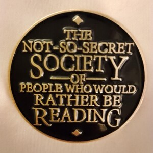 May include: A black and gold round coin with the text "THE NOT-SO-SECRET SOCIETY OF PEOPLE WHO WOULD RATHER BE READING". The coin has a gold rim and a small diamond shape at the top.