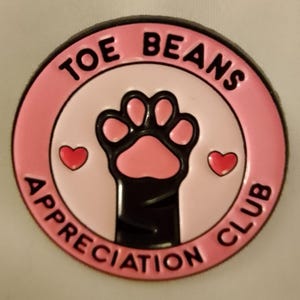 Puede incluir: Un pin redondo de esmalte rosa y negro con el texto "TOE BEANS APPRECIATION CLUB". El pin presenta una huella de pata negra con almohadillas rosas y dos pequeños corazones rojos. Un lindo accesorio para los amantes de los gatos.