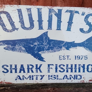 Puede incluir: Un letrero metálico rectangular con un acabado envejecido. El letrero presenta el texto "QUINT'S" sobre una ilustración de un tiburón azul. Debajo, se lee "SHARK FISHING AMITY ISLAND EST. 1975".
