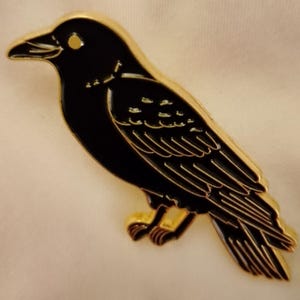 May include: A black raven brooch with gold edging. The bird is shown in profile, with detailed feather and eye markings. The brooch is likely metal and has a glossy finish.