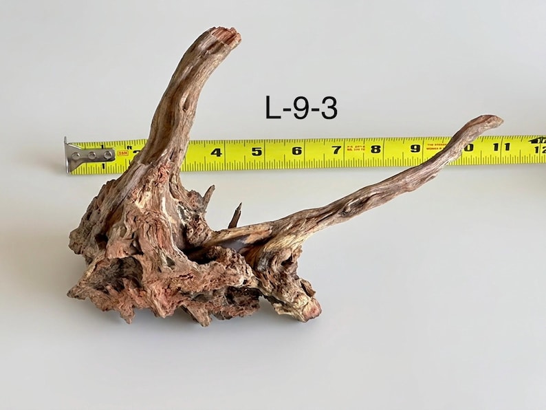 May include: A piece of brown driftwood labeled L-9-3, featuring a textured, gnarled surface with a prominent vertical branch and a long, slender horizontal extension. A yellow measuring tape shows the piece spans approximately 11 inches in length.