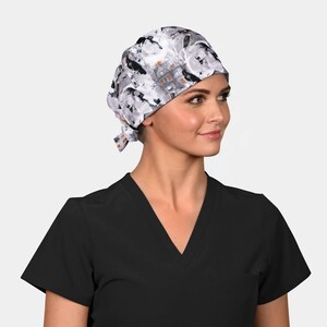 May include: A patterned surgical scrub cap featuring a Halloween theme. The cap has a white background with black bats, orange pumpkins, and a haunted house design. The cap is tied at the back and worn with a black V-neck scrub top.