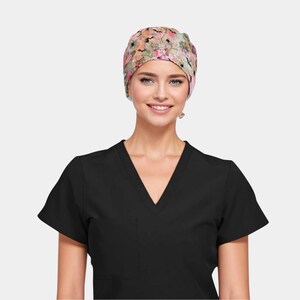 May include: A floral patterned scrub cap in shades of pink, green, and beige. The cap is worn by a person wearing a black V-neck scrub top. The cap features a wrap-around design and is made of a soft, lightweight fabric.