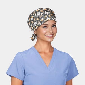 May include: A patterned surgical scrub cap featuring a repeating design of dogs in various poses. The cap is tied at the back and is worn by a person wearing a light blue V-neck scrub top.