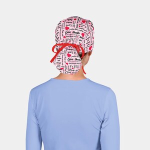 May include: A pink scrub cap with a red bow, featuring the words "Give thanks", "Love never fails", and red hearts. The cap is designed with a repeating pattern of text and hearts.