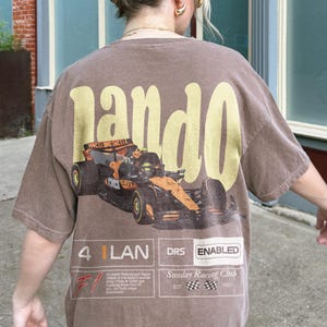 Pu&ograve; includere: T-shirt marrone con una grande grafica "Lando" gialla sul retro. Sotto, una grafica di una macchina da corsa di Formula 1 nera e arancione. Il testo aggiuntivo include "4 LAN DRS ENABLED" e "Sunday Racing Club EST 1950".