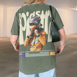 May include: Olive green t-shirt featuring a graphic of a race car driver in a helmet and orange racing suit. The shirt has the word "PIST" in a stylized font and the text "SILENT ASSASSIN" and the number "81".