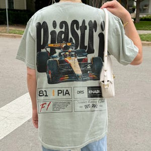 May include: Sage green t-shirt featuring a Formula 1 race car graphic with the name "Piastri" in large, distressed lettering. The design includes the number "81" and technical details. The shirt has a vintage, worn look.