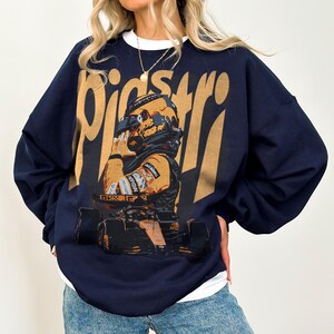 K&ouml;nnte beinhalten: Marineblaues Sweatshirt mit einem gold-orangenen Aufdruck eines Rennfahrers und Rennwagens. Das Wort "Piastri" ist in goldfarbenen, stilisierten Buchstaben aufgedruckt. Das Sweatshirt hat einen Rundhalsausschnitt und lange &Auml;rmel.