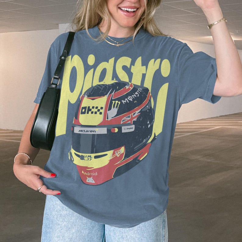 May include: A blue t-shirt featuring a graphic of a racing helmet with the name "Piastri" in yellow block letters. The helmet design includes the McLaren logo and the word "Android".