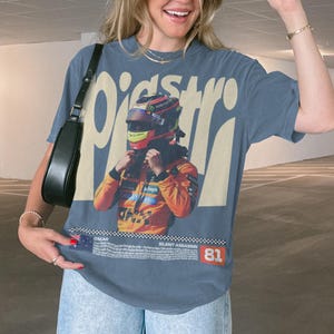 May include: Blue t-shirt featuring a race car driver in a helmet and racing suit. The shirt displays the word "Piastri" in large letters, along with the text "SILENT ASSASSIN" and the number "81".