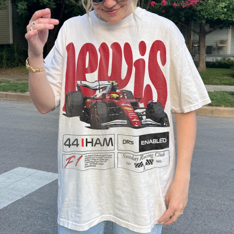 Pode incluir: Camiseta creme com um gr&aacute;fico de carro de corrida de F&oacute;rmula 1 vermelho e preto. A camisa tem a palavra "LEWIS" em letras vermelhas grandes acima do carro e texto e n&uacute;meros adicionais abaixo.