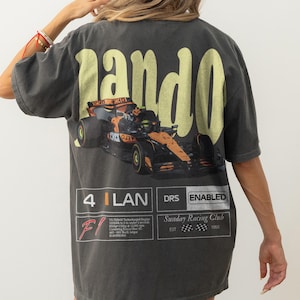 Pu&ograve; includere: T-shirt grigio scuro con un design di auto da corsa di Formula 1 e la parola "pando" in giallo. La maglietta include testo e grafica relativi alle corse, tra cui "Sunday Racing Club" e "DRS ENABLED".
