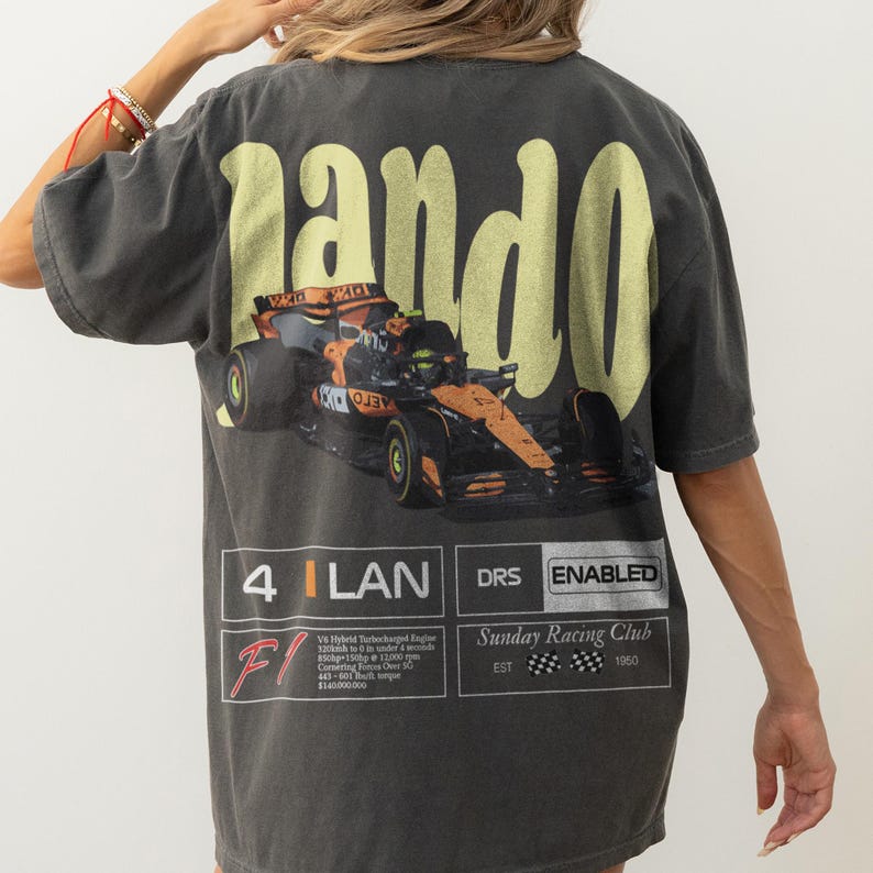 Pu&ograve; includere: T-shirt grigio scuro con un design di auto da corsa di Formula 1 e la parola "pando" in giallo. La maglietta include testo e grafica relativi alle corse, tra cui "Sunday Racing Club" e "DRS ENABLED".