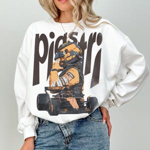 K&ouml;nnte beinhalten: Wei&szlig;es Sweatshirt mit einer Grafik eines Rennfahrers in Helm und Rennanzug, mit dem Namen "piastri" in gro&szlig;en, dunkelbraunen Buchstaben. Der Fahrer sitzt in einem Rennwagen. Das Sweatshirt hat lange &Auml;rmel.