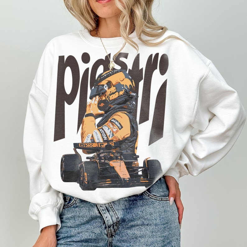 K&ouml;nnte beinhalten: Wei&szlig;es Sweatshirt mit einer Grafik eines Rennfahrers in Helm und Rennanzug, mit dem Namen "piastri" in gro&szlig;en, dunkelbraunen Buchstaben. Der Fahrer sitzt in einem Rennwagen. Das Sweatshirt hat lange &Auml;rmel.