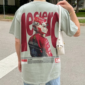 Pode incluir: Camiseta verde sálvia com um grande gráfico vermelho de um piloto de Fórmula 1 com fato e capacete vermelhos. A palavra "LECLERC" está impressa em vermelho, juntamente com o texto "PRÍNCIPE DE MÓNACO" e o número "16".