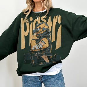 K&ouml;nnte beinhalten: Dunkelgr&uuml;nes Sweatshirt mit einem goldenen Aufdruck eines Rennfahrers und Rennwagens. Das Wort "pistar" ist goldfarben oben aufgedruckt. Der Fahrer tr&auml;gt einen Helm und einen Rennanzug. Das Sweatshirt wird mit blauen Jeans getragen.