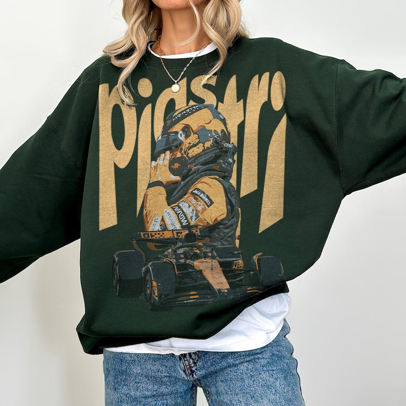 K&ouml;nnte beinhalten: Dunkelgr&uuml;nes Sweatshirt mit einem goldenen Aufdruck eines Rennfahrers und Rennwagens. Das Wort "pistar" ist goldfarben oben aufgedruckt. Der Fahrer tr&auml;gt einen Helm und einen Rennanzug. Das Sweatshirt wird mit blauen Jeans getragen.