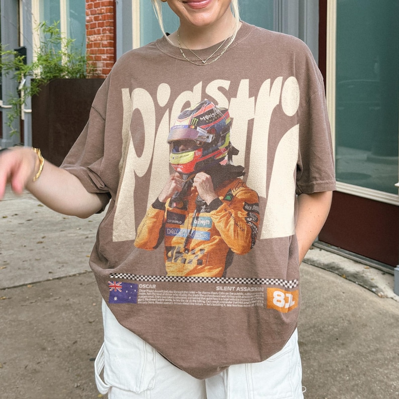 May include: A brown t-shirt with a graphic of a race car driver wearing a helmet and racing suit. The word "PISTR" is printed in large, off-white letters above the graphic. The text "OSCAR SILENT ASSASSIN 81" is printed below the graphic.
