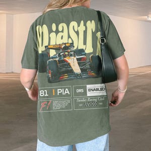 May include: Olive green t-shirt with a large graphic of a race car and the word "Piastr" in yellow. The shirt also features text that reads "81 | PIA DRS ENABLED" and "Sunday Racing Club EST 1960".