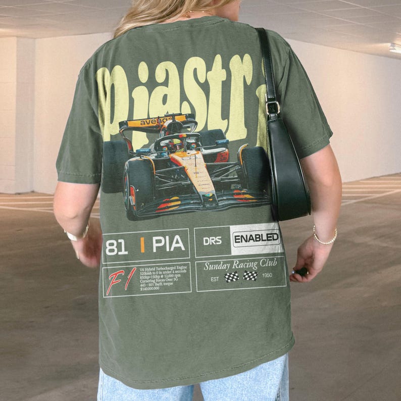 May include: Olive green t-shirt with a large graphic of a race car and the word "Piastr" in yellow. The shirt also features text that reads "81 | PIA DRS ENABLED" and "Sunday Racing Club EST 1960".