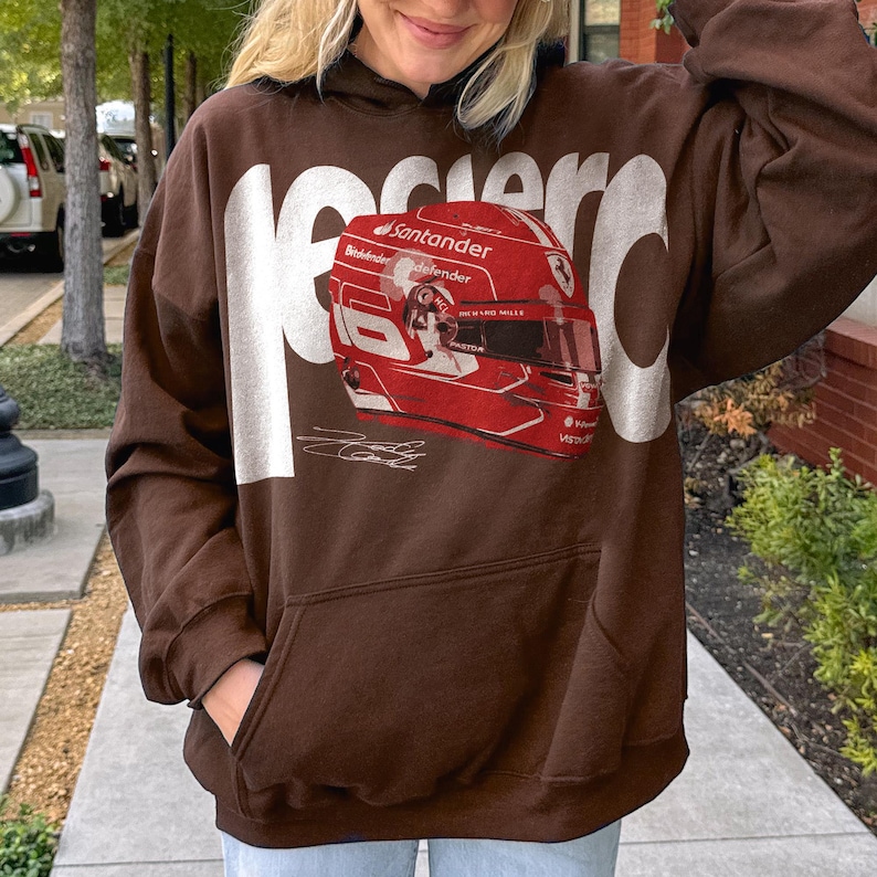 May include: Brown hoodie with a large white graphic that says "Ferrari" and a red and white racing helmet design. The hoodie has a front pocket and a drawstring hood.