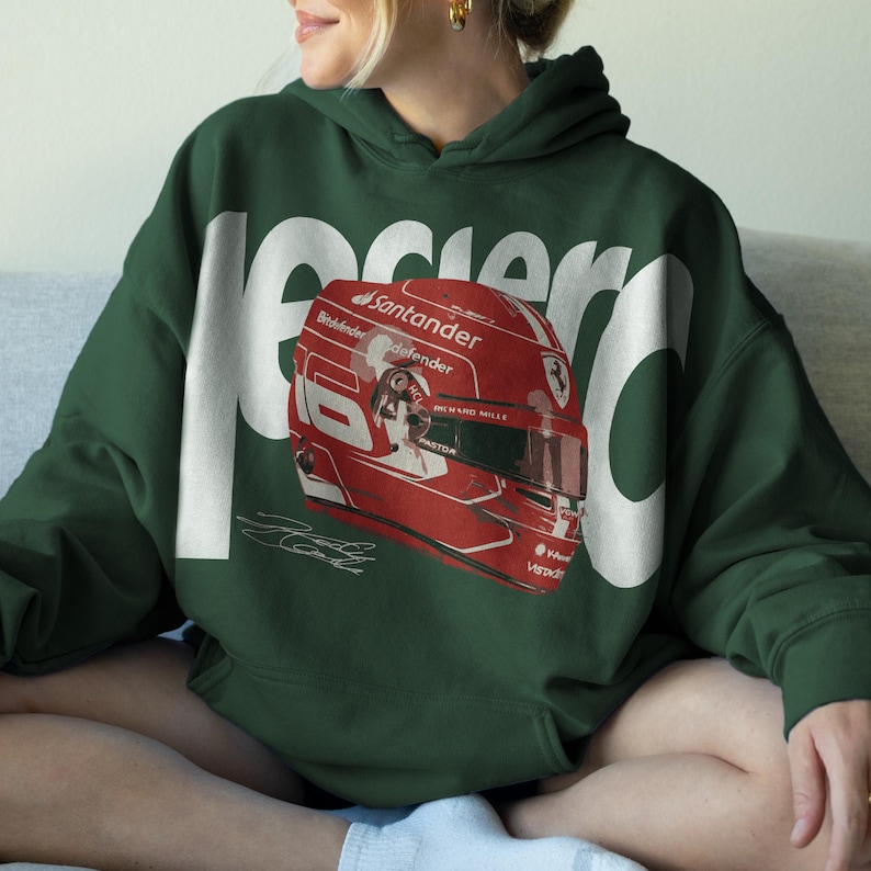 May include: Dark green hoodie featuring a large white graphic of the word "Leclerc" and a red racing helmet with the number 16. The helmet has sponsor logos and a signature. The hoodie is made of a soft material.