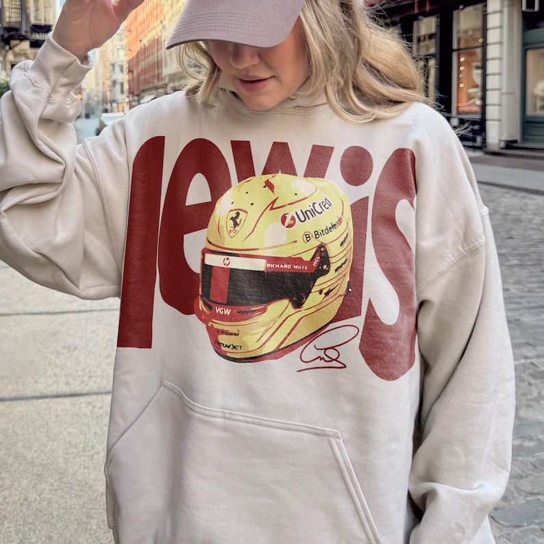May include: Beige hoodie with a large red "LEWIS" graphic and a yellow and red racing helmet design. The helmet features sponsor logos and a signature. The hoodie has a front pocket and a drawstring hood.
