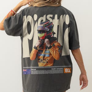 May include: Dark gray t-shirt featuring a vibrant image of a race car driver in a helmet and orange racing suit. The word "PIST" is printed on the back. The shirt also includes the text "SILENT ASSASSIN" and the number "81".