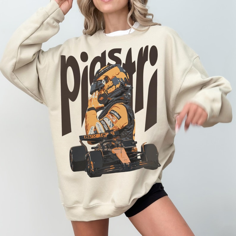 K&ouml;nnte beinhalten: Beiges Sweatshirt mit einer Grafik eines Rennfahrers mit Helm und Rennwagen. Das Wort "piastri" ist in gro&szlig;en, braunen Buchstaben &uuml;ber der Grafik aufgedruckt. Der Fahrer tr&auml;gt einen gelb-schwarzen Rennanzug.