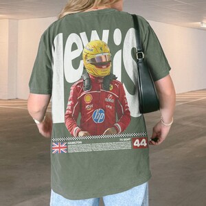 May include: Olive green t-shirt with a graphic of a Formula 1 driver in a red racing suit and yellow helmet. The text "LEWIS" is printed in white, with the number "44" and the words "7X GOAT" also visible.