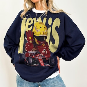 Peut inclure: Sweat-shirt bleu marine avec un motif d'un pilote de Formule 1 portant un casque jaune et une combinaison rouge, avec le nom "Lewis" en grandes lettres jaunes. Le pilote est assis dans une voiture de course rouge.