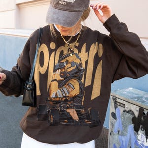 K&ouml;nnte beinhalten: Ein braunes Sweatshirt mit einem goldenen Aufdruck eines Rennfahrers und Rennwagens. Dar&uuml;ber steht das Wort "piceri" in Gold. Die Person tr&auml;gt eine graue Kappe und goldene Halsketten. Ein l&auml;ssiges Kleidungsst&uuml;ck.