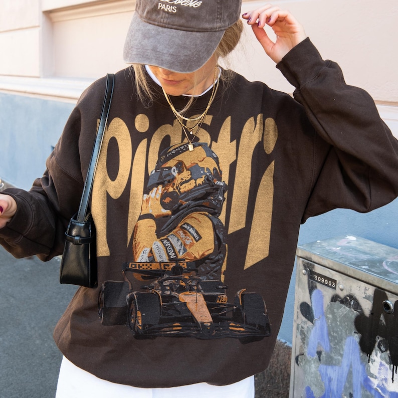 K&ouml;nnte beinhalten: Ein braunes Sweatshirt mit einem goldenen Aufdruck eines Rennfahrers und Rennwagens. Dar&uuml;ber steht das Wort "piceri" in Gold. Die Person tr&auml;gt eine graue Kappe und goldene Halsketten. Ein l&auml;ssiges Kleidungsst&uuml;ck.