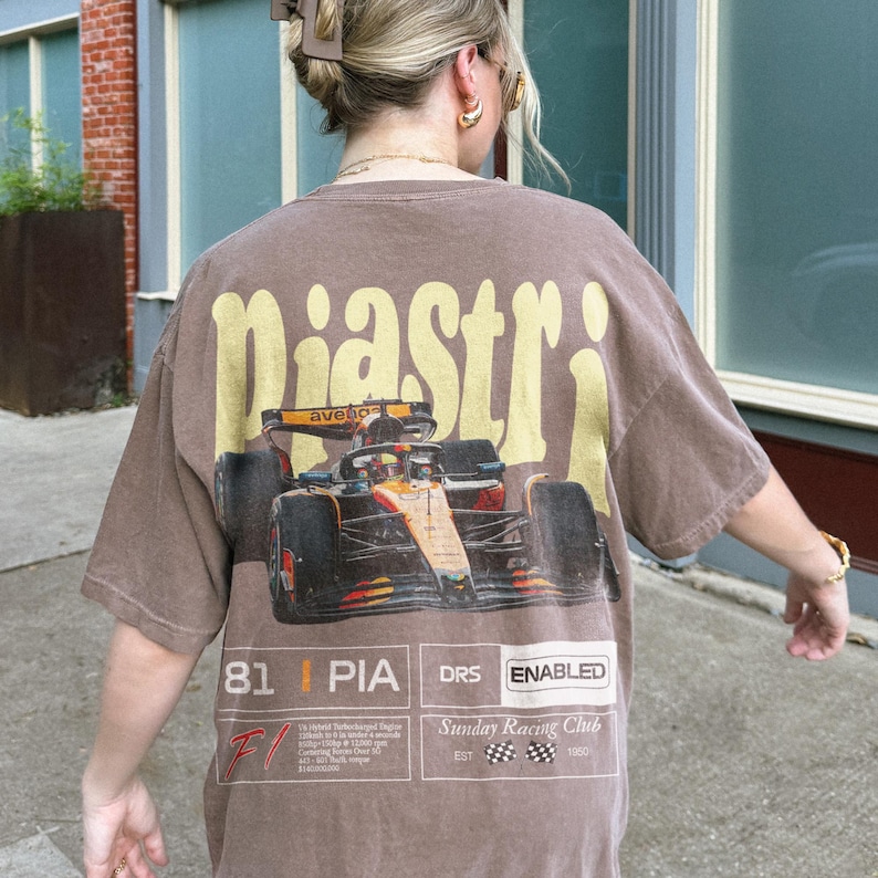 May include: Brown t-shirt with a large graphic of a race car and the name "Piastri" in yellow lettering across the back. Additional text includes "81 PIA DRS ENABLED" and "Sunday Racing Club".