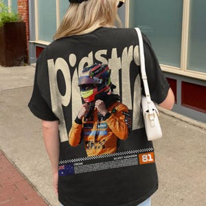 May include: Black t-shirt featuring a graphic of a race car driver in a helmet and orange racing suit. The shirt has the word "PISTONS" printed on the back in a cream color. The text "SILENT ASSASSIN" and the number "81" are also visible.