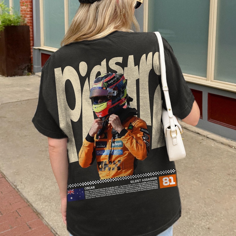 May include: Black t-shirt featuring a graphic of a race car driver in a helmet and orange racing suit. The shirt has the word "PISTONS" printed on the back in a cream color. The text "SILENT ASSASSIN" and the number "81" are also visible.