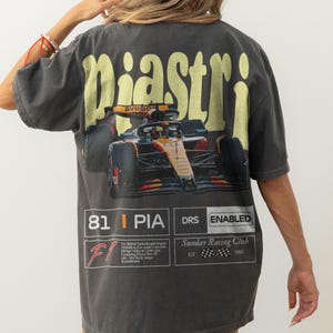 May include: Dark gray t-shirt featuring a Formula 1 race car graphic with the name "piastri" in yellow arched text. The shirt includes racing details and the text "Sunday Racing Club".