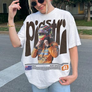 May include: White t-shirt with a graphic of a race car driver in a helmet and orange racing suit. The word "piastri" is printed in large brown letters above the image. The shirt also includes the text "OSCAR" and "SILENT ASSASSIN" with the number "81".