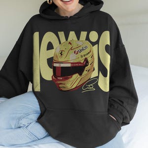 May include: Black hoodie with the name "LEWIS" in gold block letters. The hoodie features a graphic of a yellow and red racing helmet with sponsor logos and a signature. The helmet design includes a red stripe and a black visor.