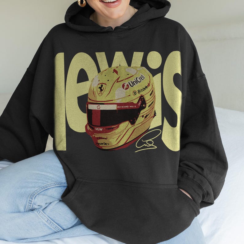 May include: Black hoodie with the name "LEWIS" in gold block letters. The hoodie features a graphic of a yellow and red racing helmet with sponsor logos and a signature. The helmet design includes a red stripe and a black visor.