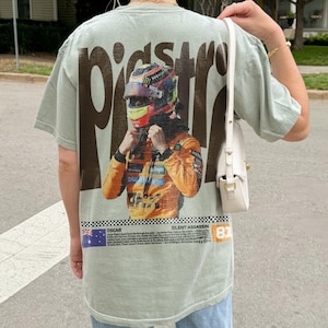 May include: Sage green t-shirt featuring a graphic of a race car driver in a helmet and racing suit. The word "PISTONS" is printed in large brown letters above the graphic. The bottom of the shirt has text and the number 81.